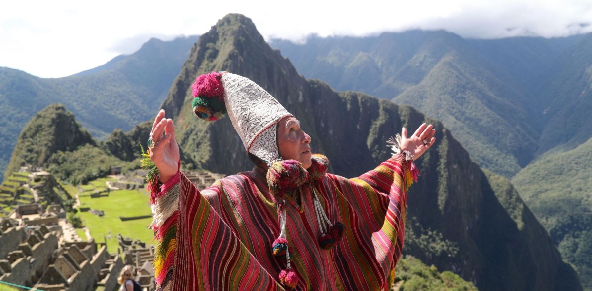 Hombre peruano vestido como los antiguos chamanes con los brazos en alto mirando al cielo, detrás la ciudad antigua de Machu Pichu
