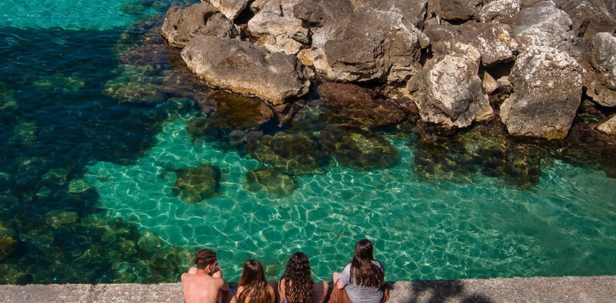 tres chicas y un chico de espalda sentados en una toalla mirando al mar
