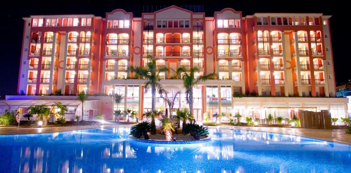 Fachada de un hotel con una piscina al frente y en el medio unas palmeras