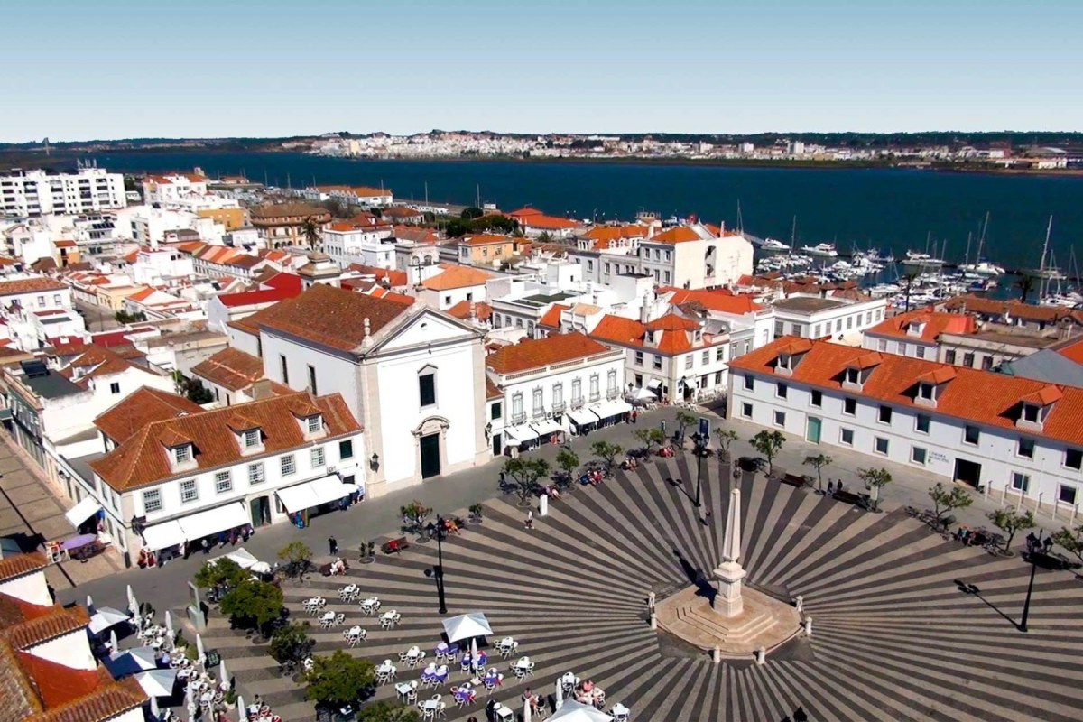 Plaza de un pueblo rodeada de casas de color blanco, al fondo un rio