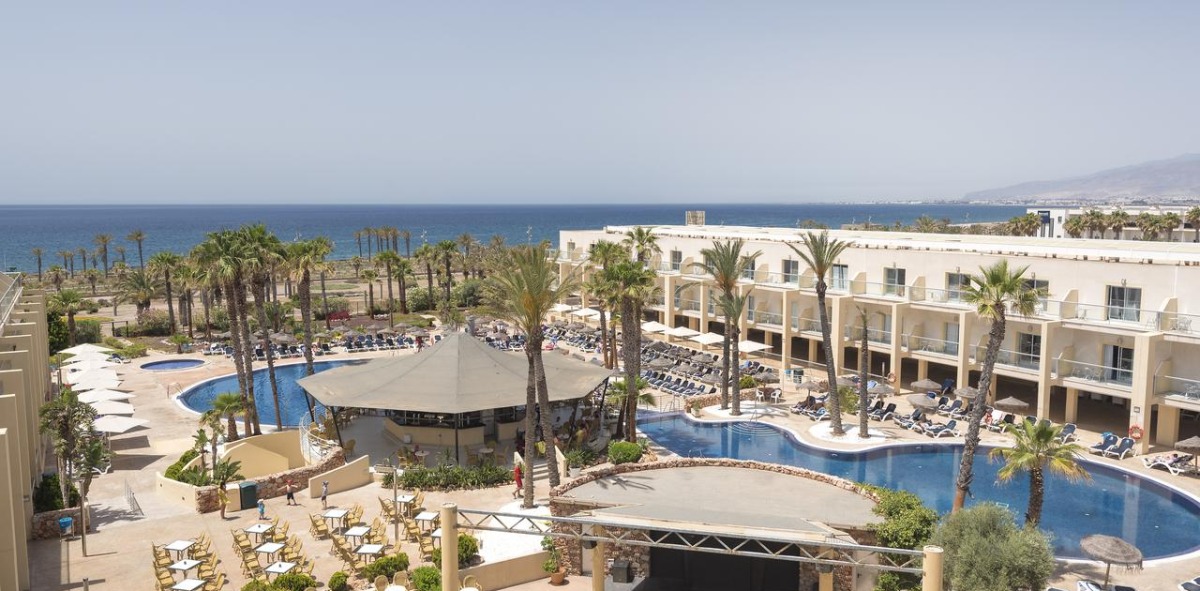 Piscina de un hotel de color blanco con palmeras y al fondo el mar