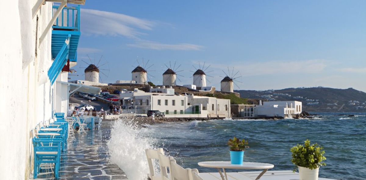 Una terraza con mesas y sillas al lado del mar y los famosos molinos de Mikonos al fondo