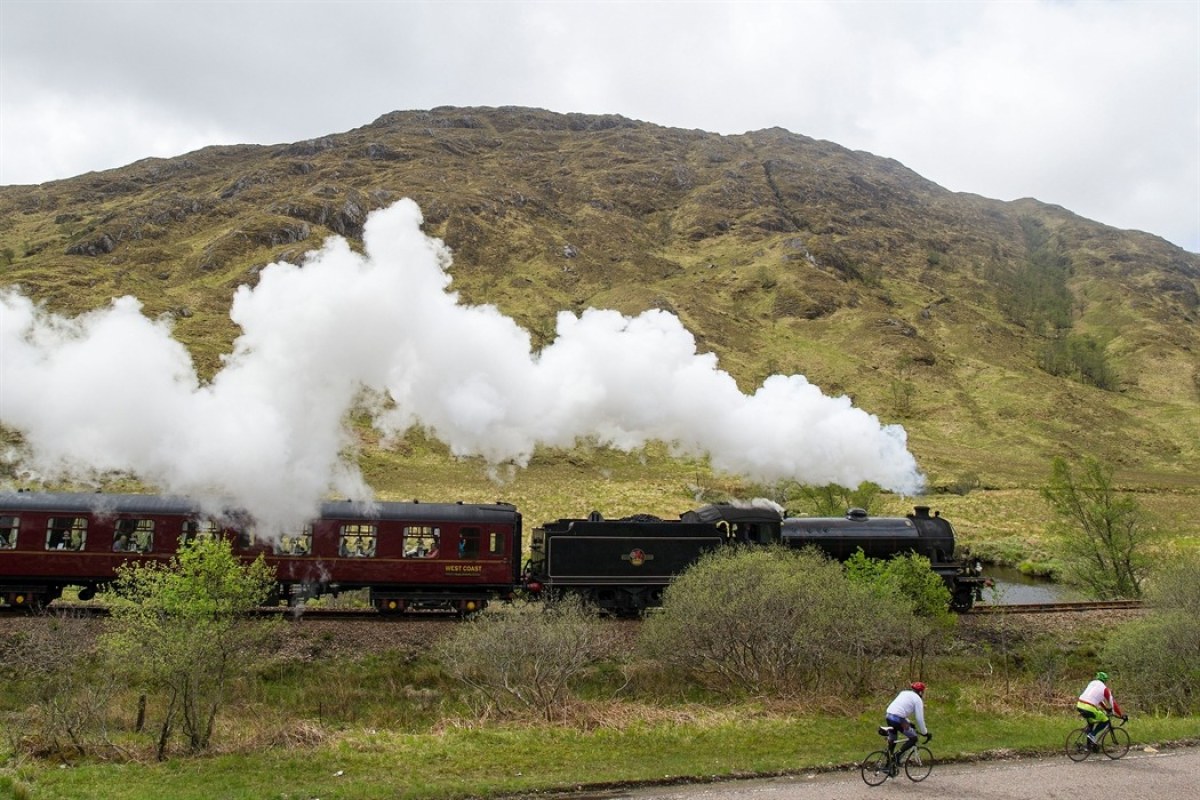 Tren de vapor por un paisaje verde y a su lado dos personas en bicis