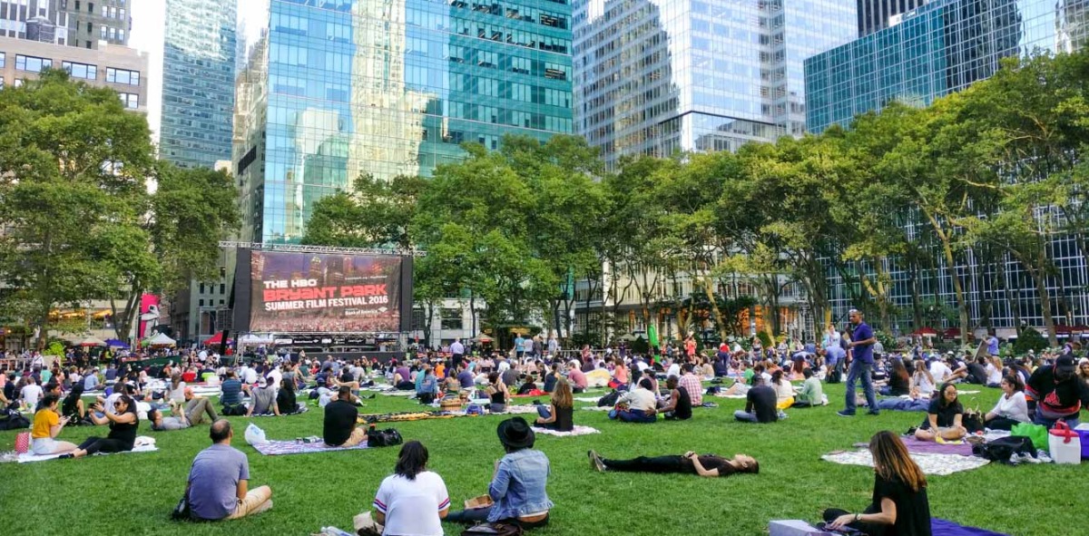 Parque de hierba lleno de gente sentada y tumbada, al fondo un escenario y rodeado de edificios