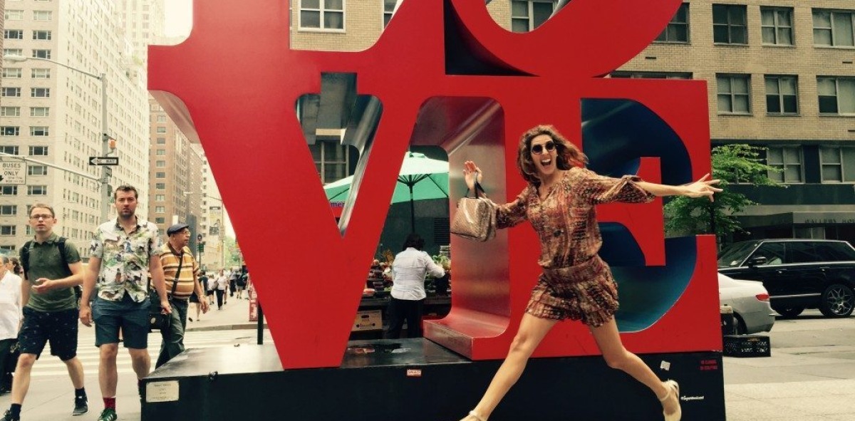 Mujer saltando delante de una estatua que pone Love en una calle con mucha gente mirando
