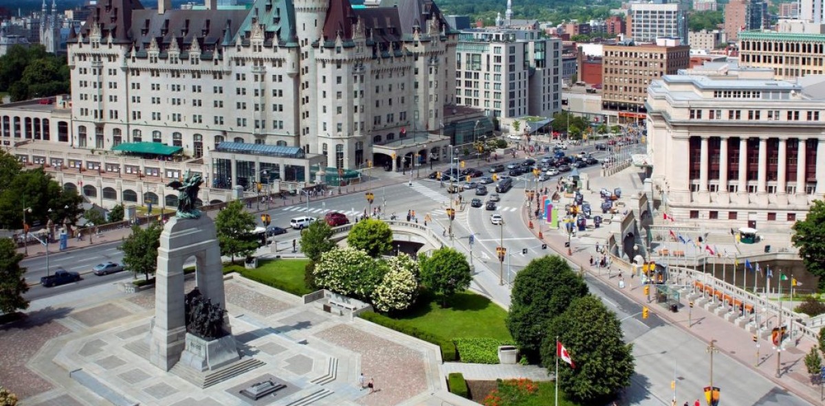 Panorámica de una plaza de Ottawa donde aparece en el medio de la plaza un monumento y un gran edificio con torreones