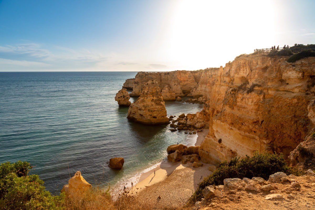 vista desde lo alto de rocas entrando al mar junto a una playa donde se ven personas paseando