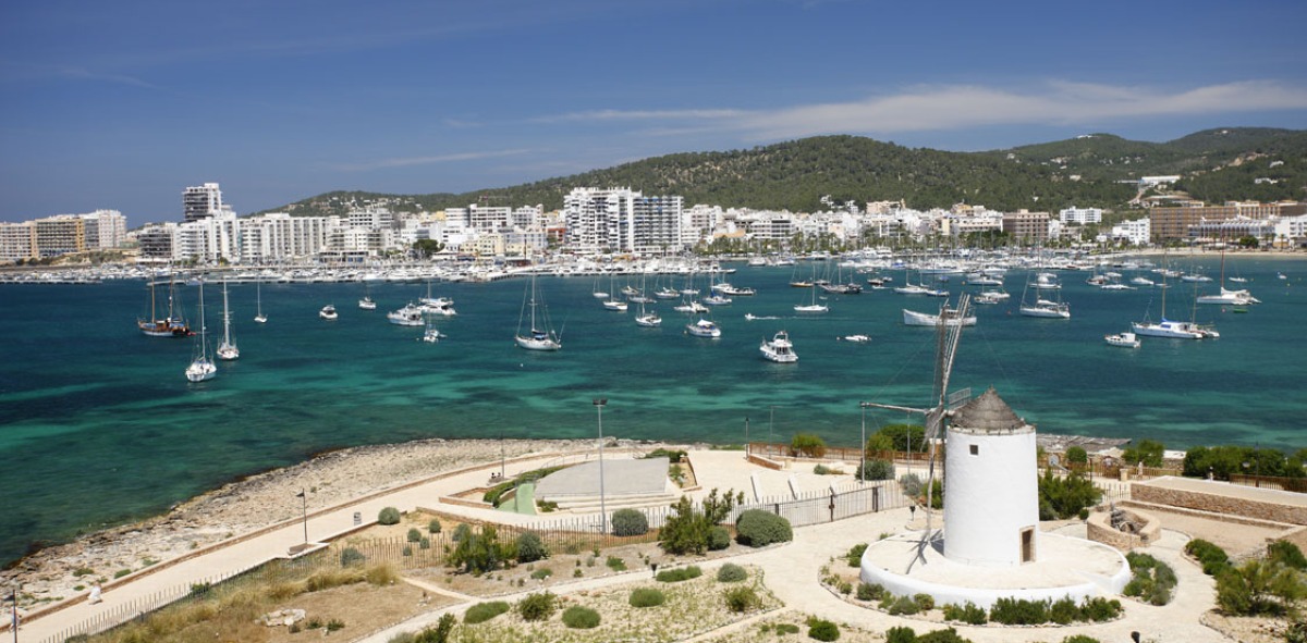 Panorámica de Ibiza al fondo con barcos atracados y un molino de viento
