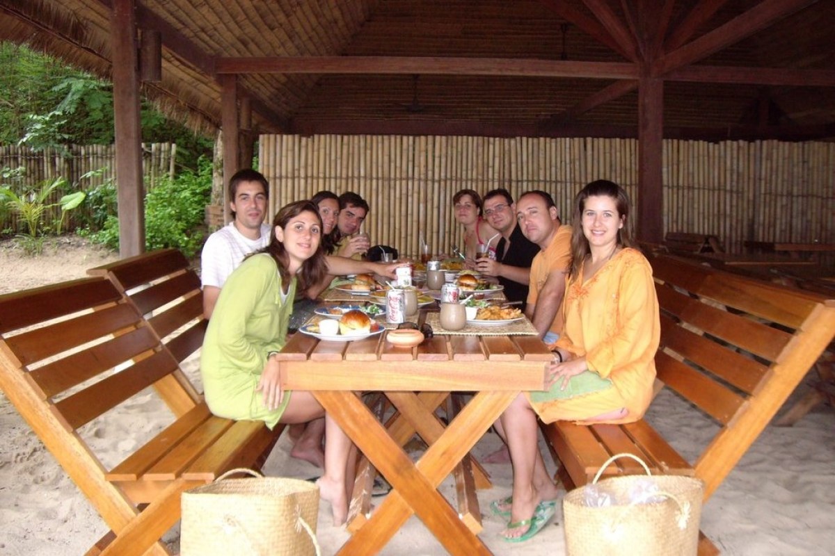 Grupo de solteros viajeros sentados en bancos de madera y mesa de madera mirando a la persona que hace la foto y disfrutando de una comida