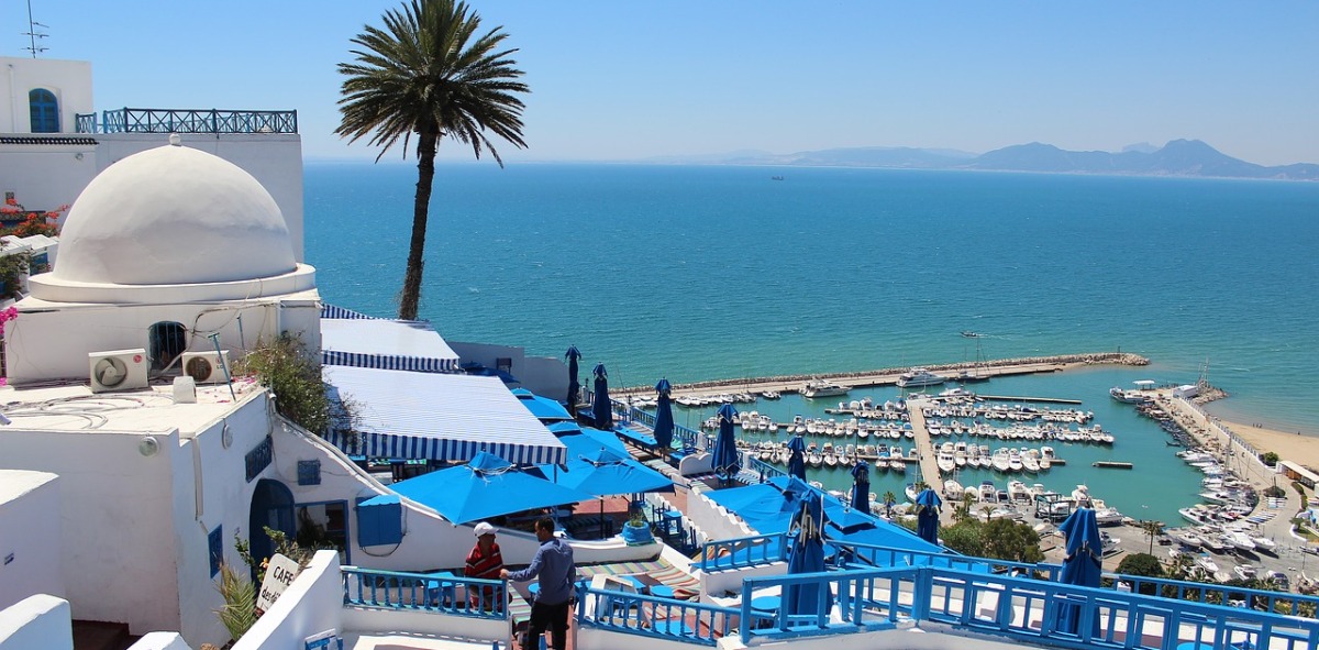 En primer plano casas blancas con toldos azules y al fondo puerto marítimo rodeado del mar azul