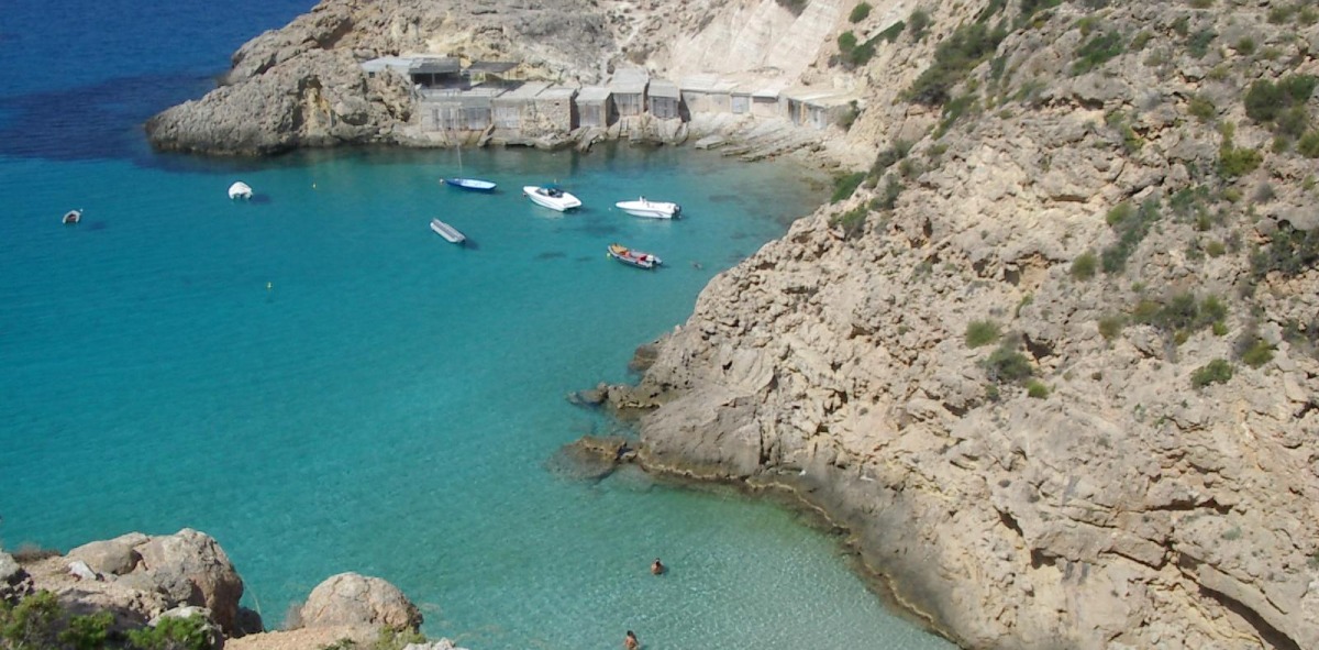 Cala de Ibiza con gente bañándose en aguas cristalinas y con barquitas atracadas