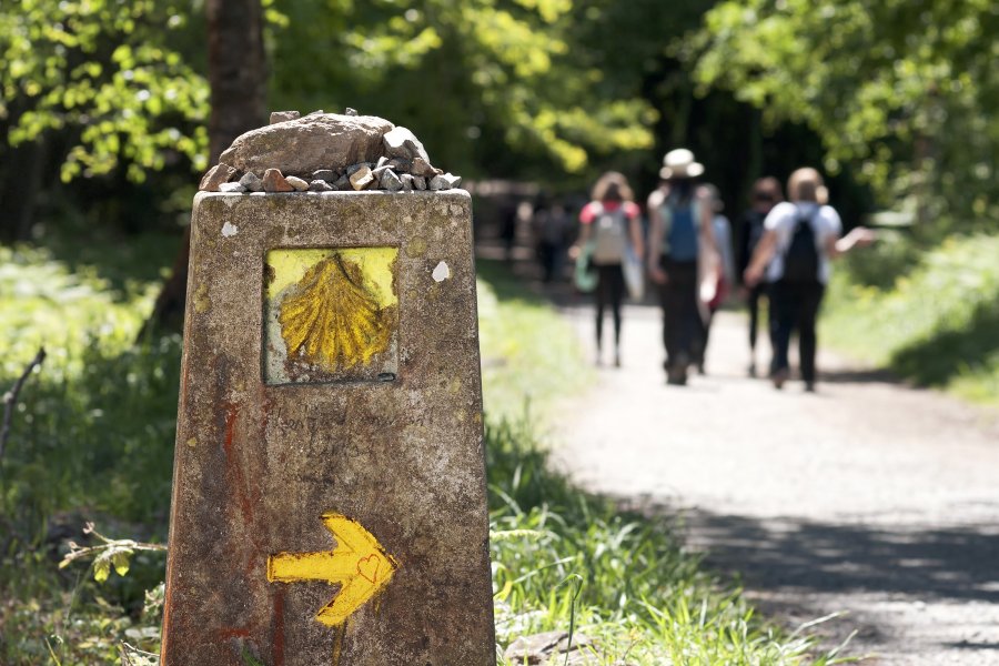 Señalización en piedra con una flecha y cocha amarilla señalando el camino, al fondo grupo de peregrinos