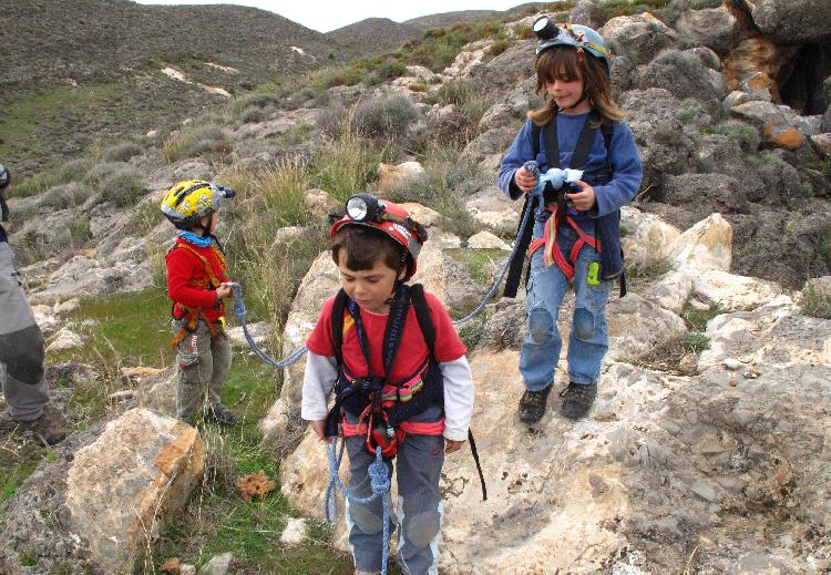 Tres niños en el monte, vestidos con ropa de senderismo, cascos y focos subidos en piedras