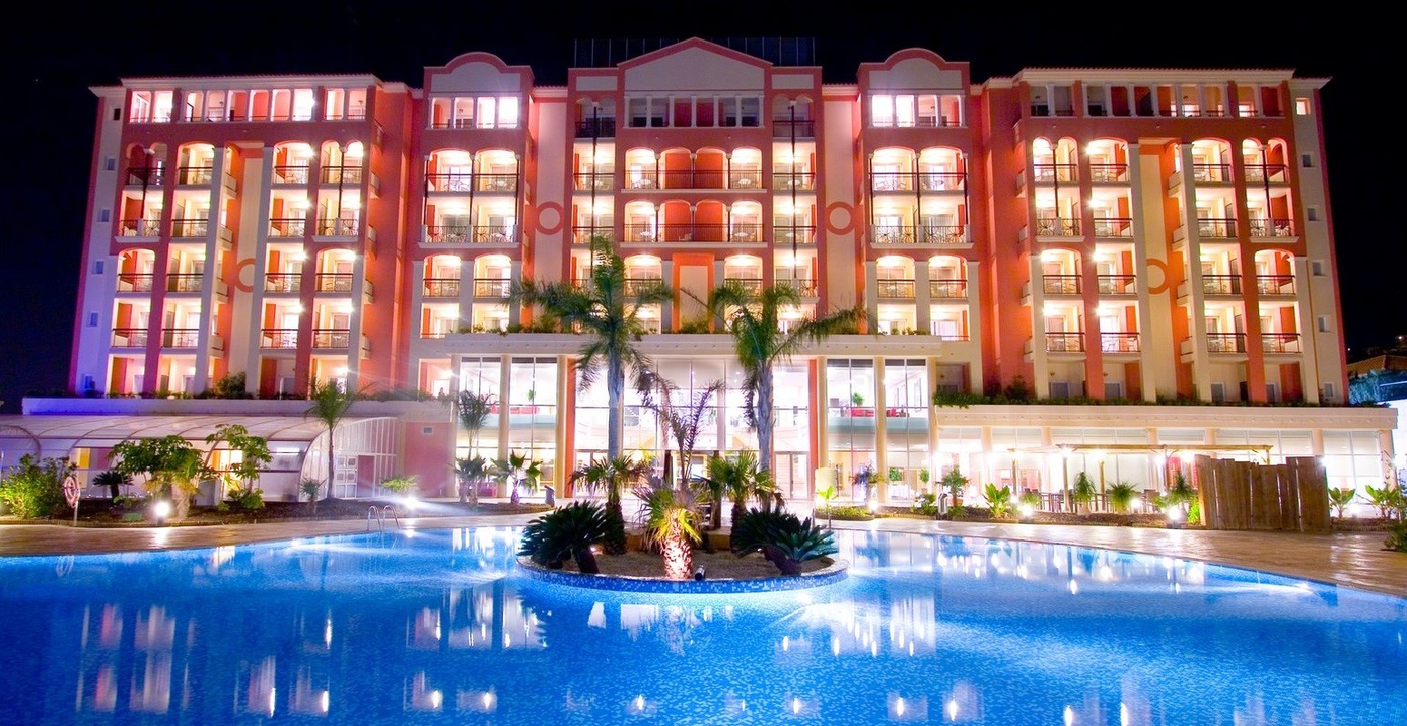 Fachada de un hotel con una piscina al frente y en el medio unas palmeras