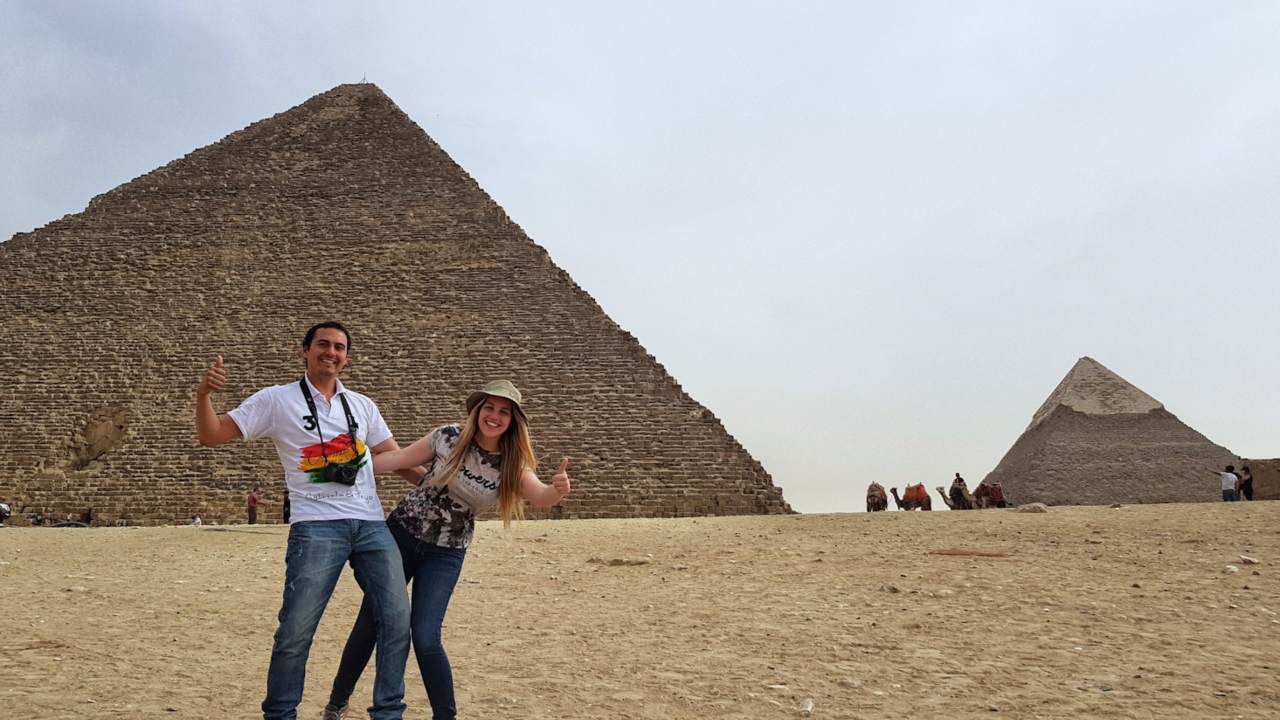 Chico y chica posando antes la pirámide de Giza, al fondo la pirámide de Keops y unos camellos paseando