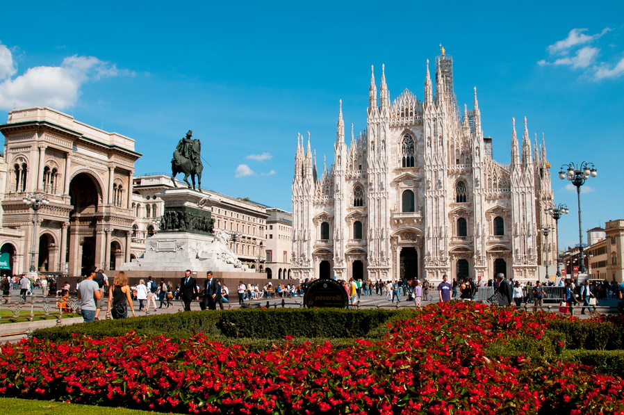 Panorámica de la catedral de Milán en una plaza con muchas flores rojas y una estatua