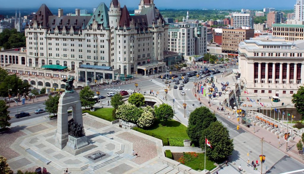 Panorámica de una plaza de Ottawa donde aparece en el medio de la plaza un monumento y un gran edificio con torreones