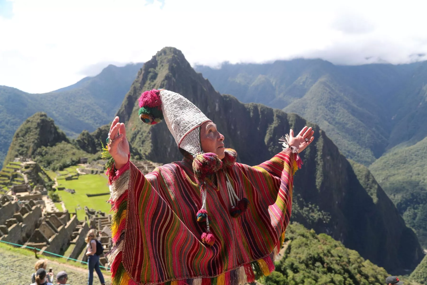 Hombre peruano vestido como los antiguos chamanes con los brazos en alto mirando al cielo, detrás la ciudad antigua de Machu Pichu