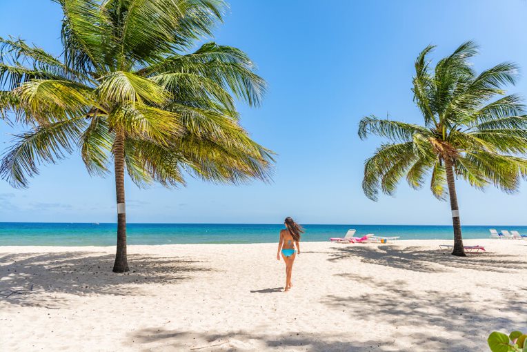 Mujer de espaldas con un biquini azul paseando por la playa de arenas blanca y dos palmeras