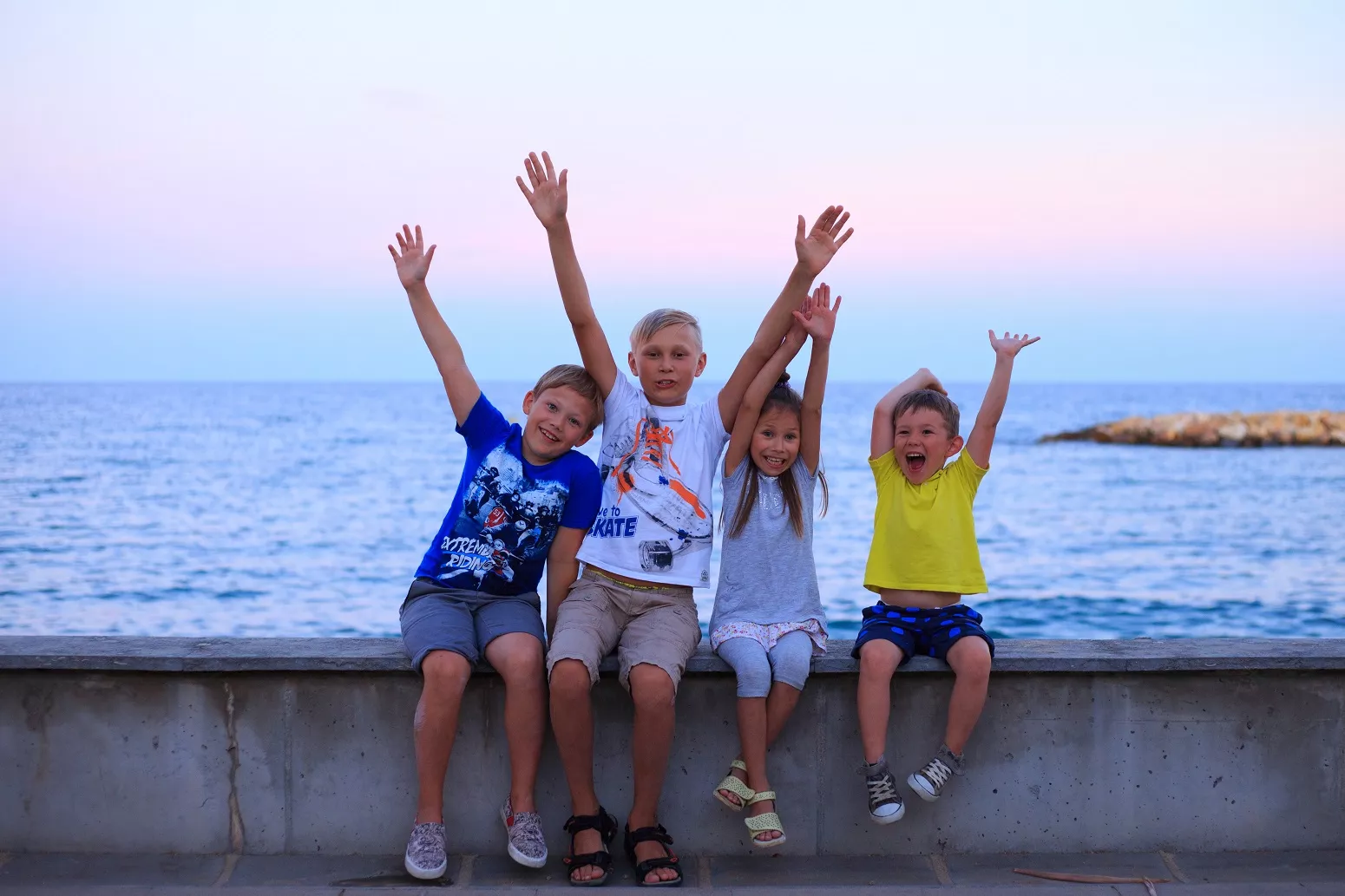 Cuatro niños sentados al borde del mar con los brazo alzados saludando