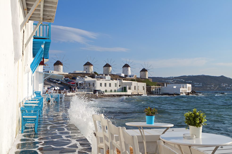 Una terraza con mesas y sillas al lado del mar y los famosos molinos de Mikonos al fondo