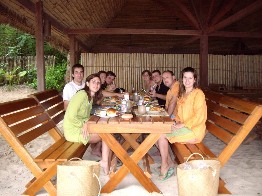Grupo de solteros viajeros sentados en bancos de madera y mesa de madera mirando a la persona que hace la foto y disfrutando de una comida